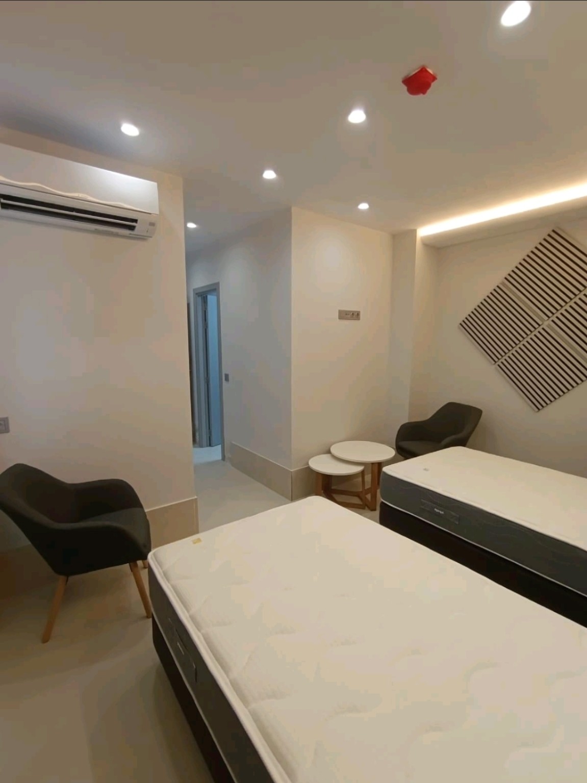 Detalle de habitación con dos camas, butacas oscuras y mesas auxiliares blancas